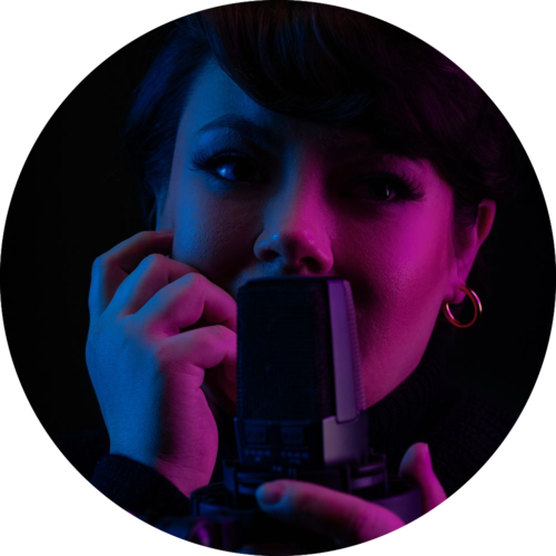 Sydnee Fullmer in a stylistic photo, under shadowy purple lighting, holding a microphone in front of her, with her other hand by her face.