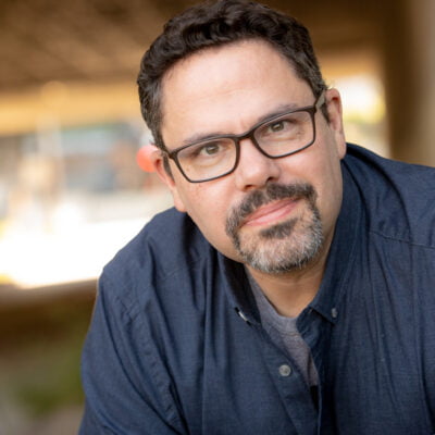 Jeremy Hall: White man with dark hair, goatee with some gray, wearing glasses, and a blue button down shirt with a gray undershirt, in front of an outdoor background that is not in focus.