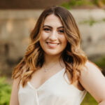 This photo is of Jess Giusti, our Media Marketing Manager. She's a white woman (Italian heritage), smiling, she has brown hair that is highlighted and she's wearing a pretty white dress.