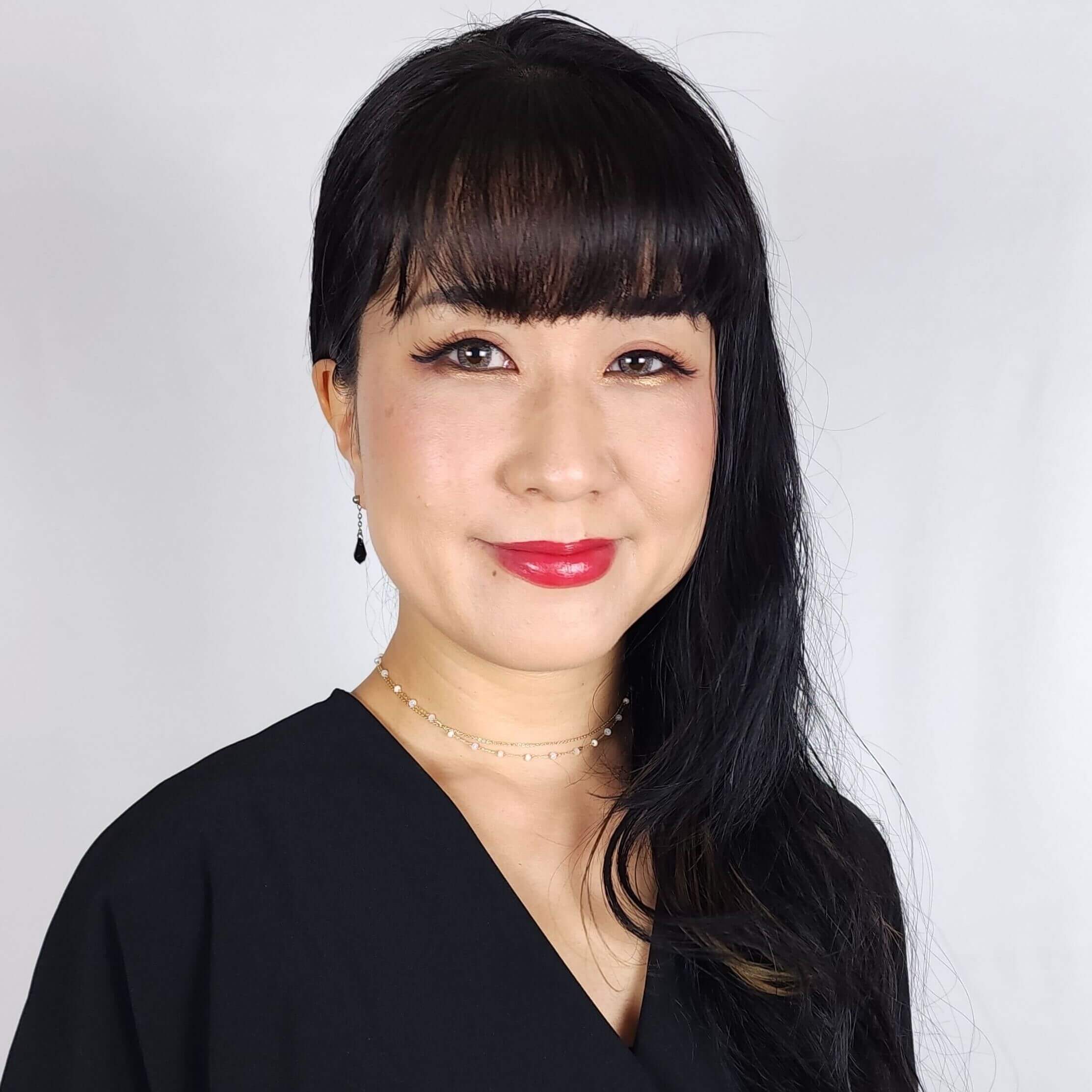 Anime Casting Director Mami Okada. Asian woman wearing a black top with black hair laying on her left shoulder, smiling wearing red lipstick.