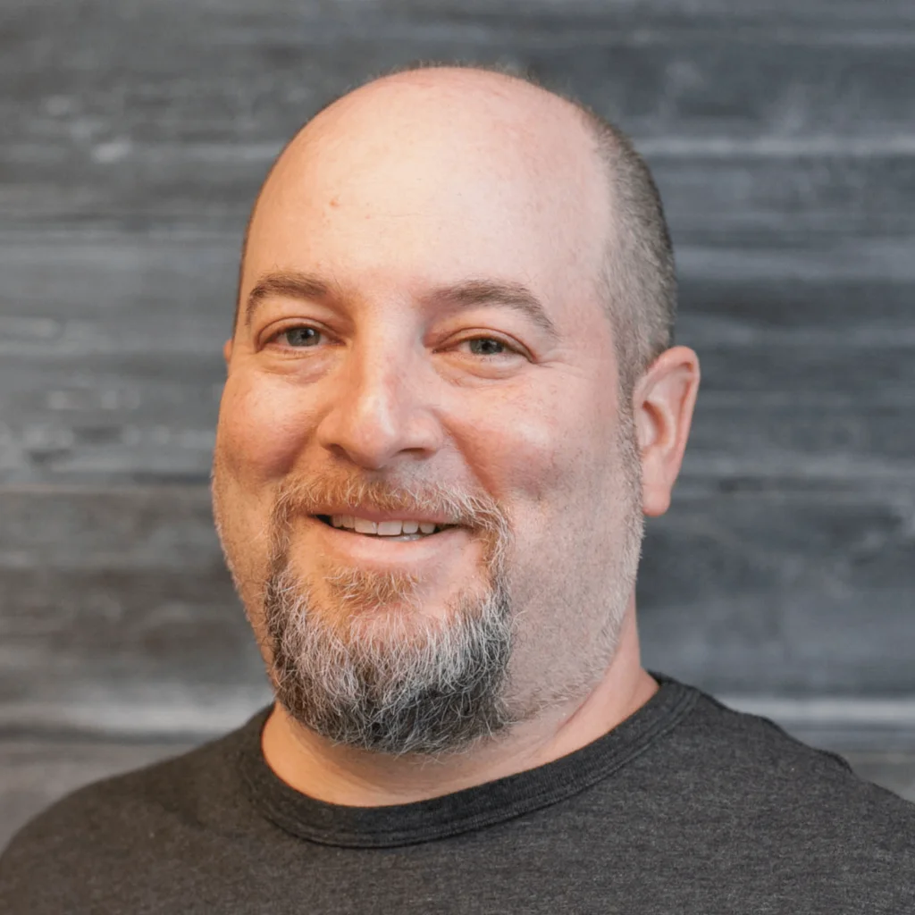 A white man with short shaved hair and a goatee smiling wearing a brown crew neck tshirt.